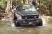 1991 Ford Bronco - A black pickup truck navigates through a rocky stream, kicking up water as it goes.