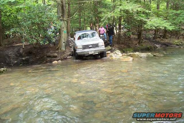Silver 1991 Ford Bronco driving through a shallow stream in a wooded area