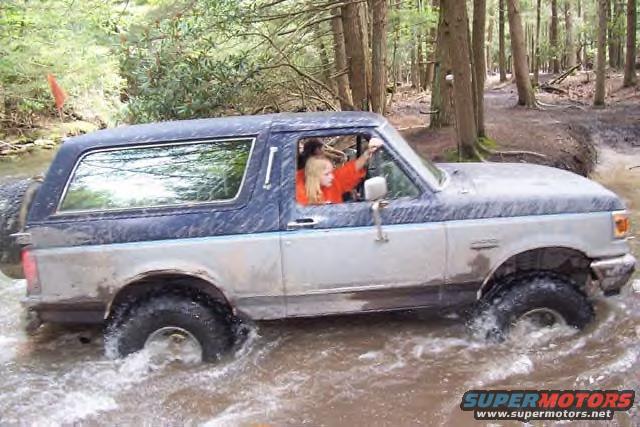 Two-tone blue and silver Ford Bronco driving through shallow water in a forest