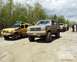 1991 Ford Bronco - A line of off-road vehicles, including a gray Ford Bronco, are parked on a dirt path with trees in the background.