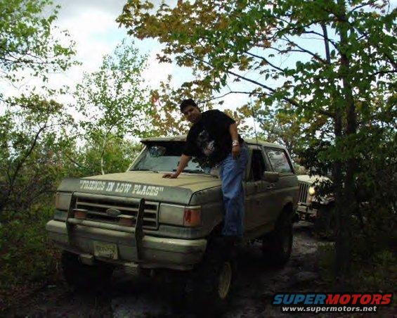 1987 Ford Bronco with "FRIENDS IN LOW PLACES" on hood, person leaning on it. Another Jeep visible behind