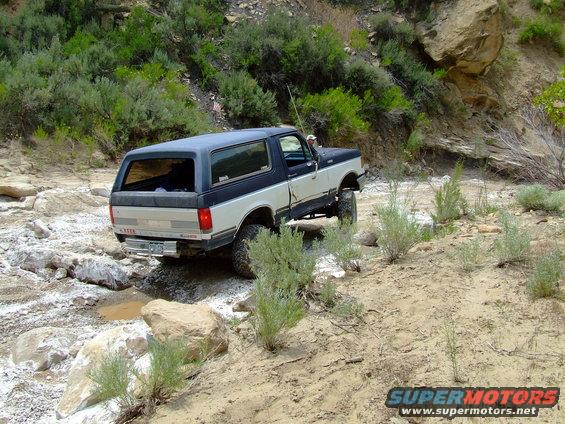 Two-tone blue and silver 1987 Ford Bronco off-roading on a rocky, dry creek bed