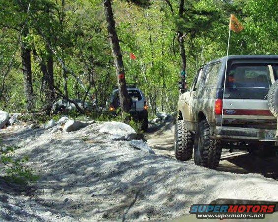 Two Broncos driving on a dirt trail through trees. The one in the foreground is a 1991 Ford Bronco