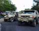 1991 Ford Bronco - Two off-road vehicles, a Ford Bronco and a Jeep CJ, are parked side-by-side on a dirt path with trees in the background.