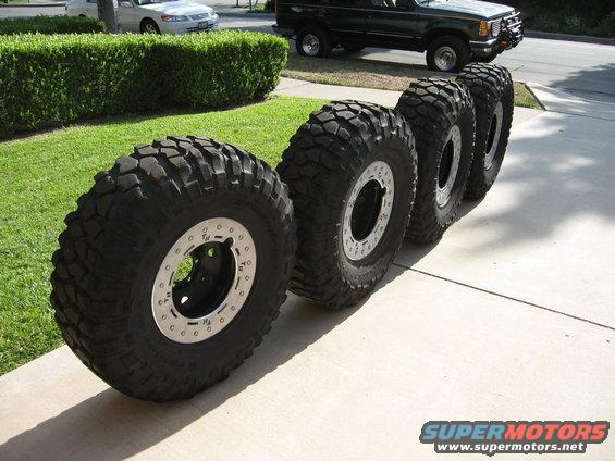 Four large off-road tires with silver beadlock rings lined up on a concrete driveway