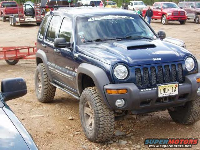 Blue Jeep Liberty with off-road tires parked on dirt. Other vehicles and trailers in background