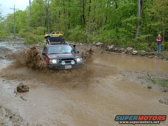 Red SUV splashing through a muddy puddle on a dirt road, with a yellow SUV behind it