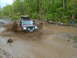1991 Ford Bronco - A red SUV navigates a deep mud puddle on a wooded trail, with another vehicle following.