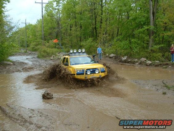 Yellow SUV with roof lights splashes through mud puddle on a dirt road