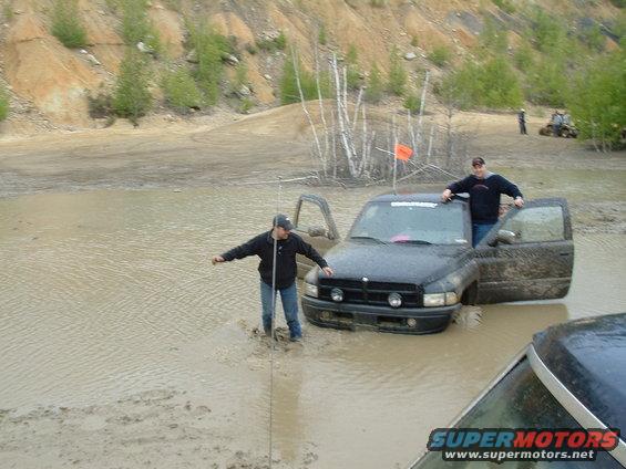 Black Dodge Ram pickup truck stuck in mud with two guys standing around it. Another vehicle is partially visible in the foreground