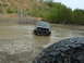 1991 Ford Bronco - A black Dodge Ram truck is being pulled through a muddy water obstacle, with other off-road vehicles visible in the distance.