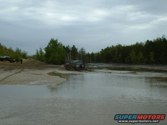 Off-road vehicles navigating a muddy, watery terrain with trees in the background