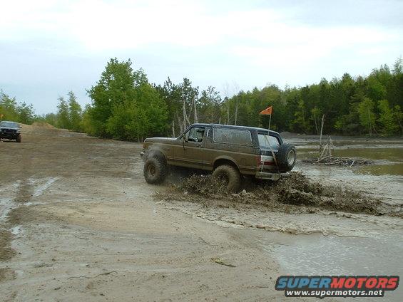 Muddy 1991 Ford Bronco driving through a dirt and water obstacle course. Another SUV is visible in the background