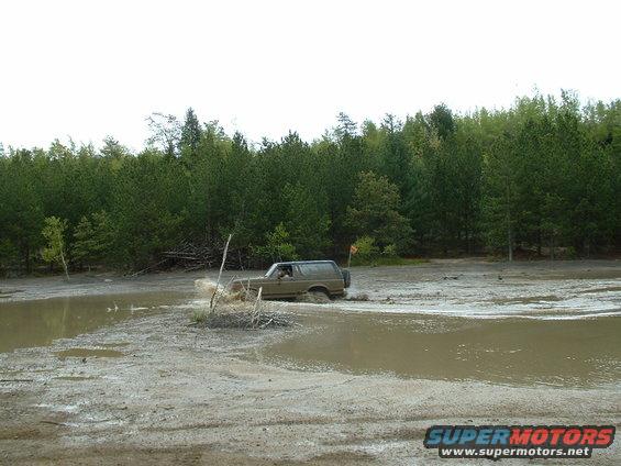 1991 Ford Bronco driving through a muddy area with trees in the background
