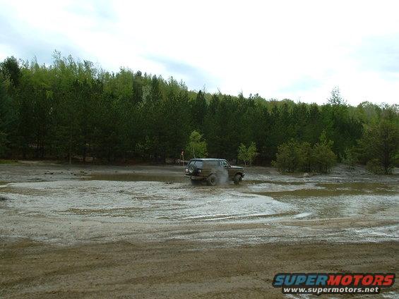 1991 Ford Bronco driving through a muddy field with trees in the background