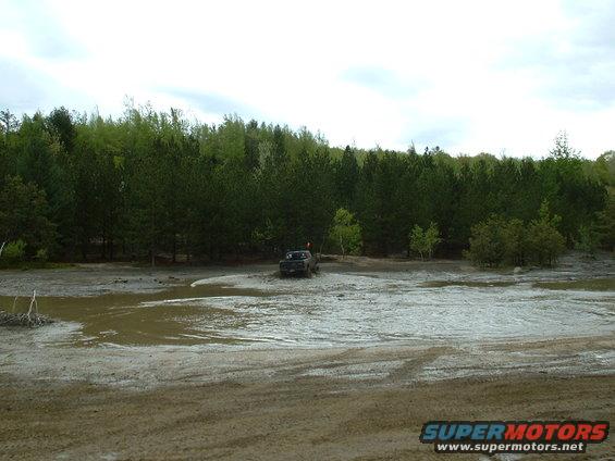 Dark colored truck driving through a muddy, wet area with trees in the background