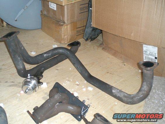Exhaust manifold and pipes with a turbocharger wastegate, on a wooden floor with cardboard boxes in the background