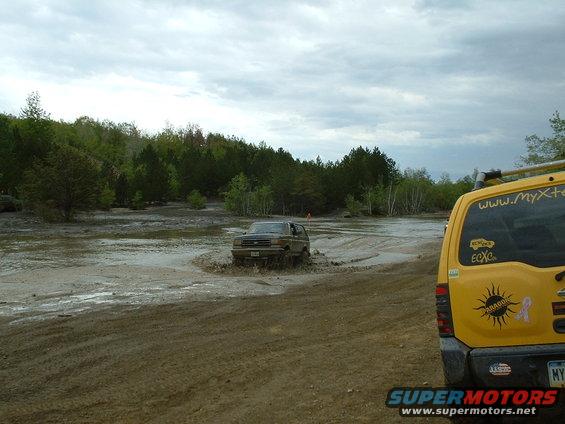 Brown SUV driving through mud puddle, with a yellow SUV in the foreground