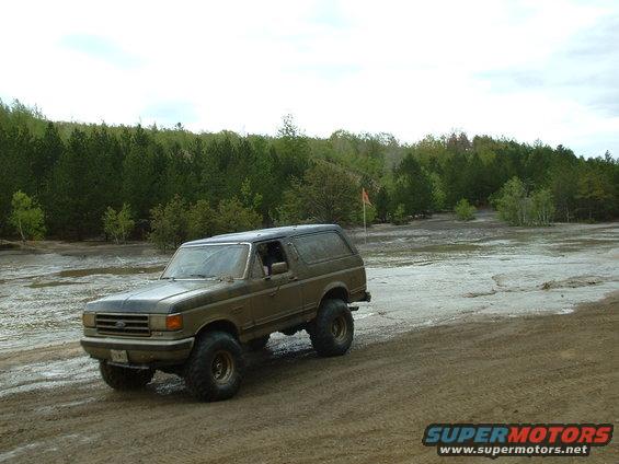 1991 Ford Bronco driving on a muddy trail with trees in the background