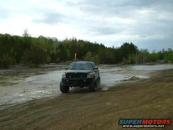 Black pickup truck with large tires driving through mud and water, trees in background