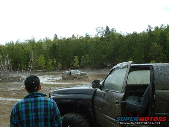 Two trucks driving through a muddy area with trees in the background