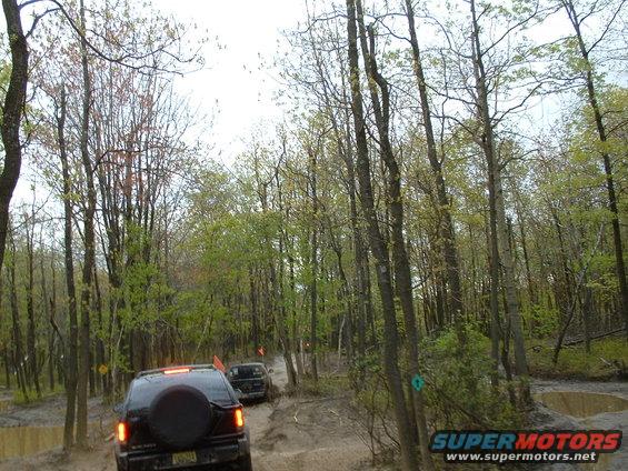 Black SUV with spare tire on rear door driving on muddy trail through trees