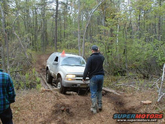 Silver SUV with an orange flag on the windshield navigates a rough dirt path in a wooded area