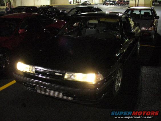 Front view of a black 1991 Mazda 626 parked at night, with other cars around