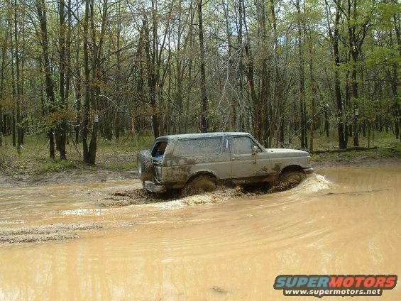 Mud-covered 1987 Ford Bronco driving through a muddy puddle in a wooded area