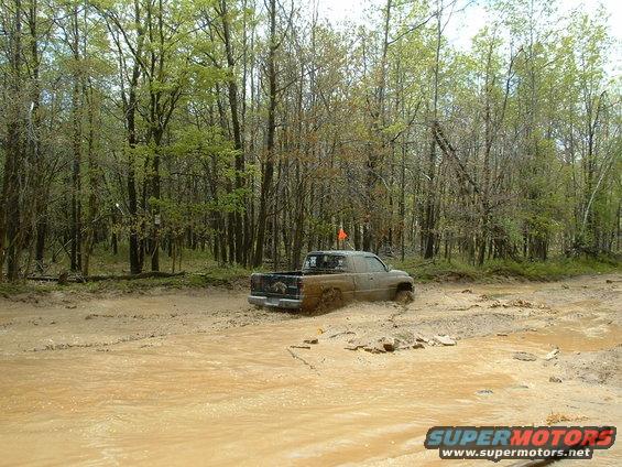 Truck stuck in a muddy path with trees on both sides. Orange flag on antenna