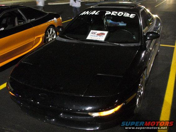 Black coupe with 'ANAL PROBE' sticker on windshield, parked next to an orange and black car