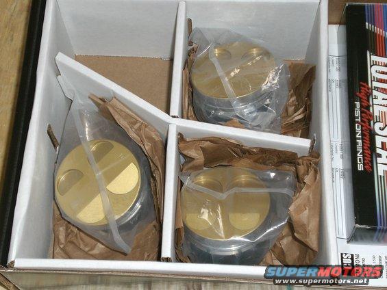 Four gold pistons in plastic bags inside a white cardboard box with a box of piston rings to the right