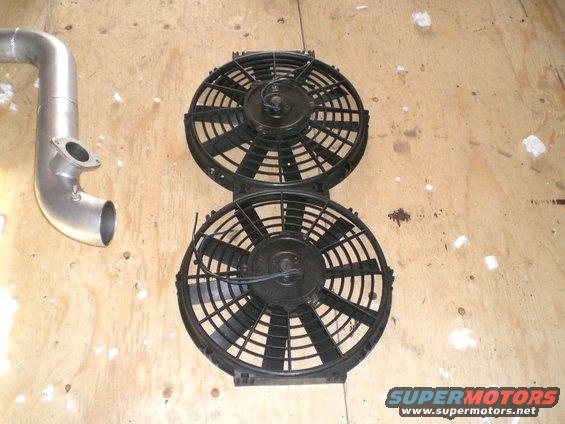 Two black electric fans stacked vertically and a silver metal pipe with a flange on a wooden surface