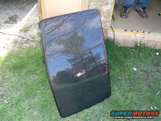 Black car sunroof panel lying on grass next to a wooden structure