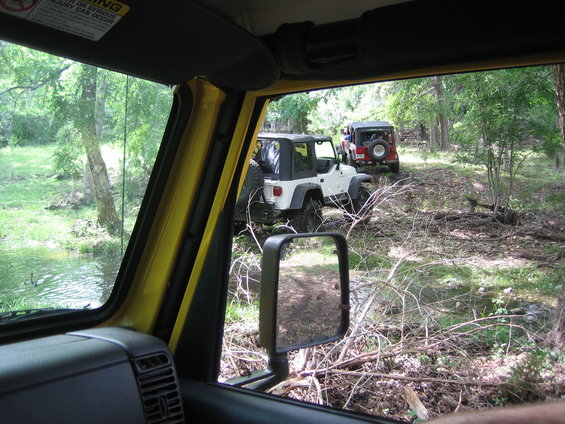 Two Jeep Wranglers driving on a dirt trail through a wooded area with a stream on the left