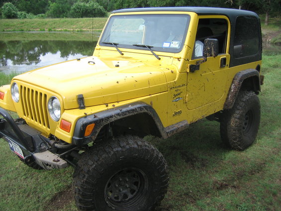 Yellow 2001 Jeep Wrangler with black hardtop, muddy tires, and aftermarket front bumper parked near a pond
