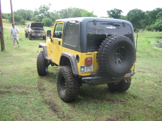 Yellow 2001 Jeep Wrangler seen from the rear, with a spare tire mounted. Another truck is visible in the background