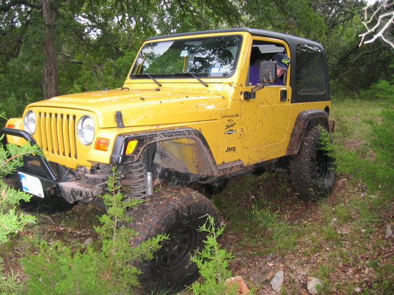 Yellow 2001 Jeep Wrangler with black hardtop, off-road tires, and aftermarket bumper, parked on a dirt path surrounded by trees