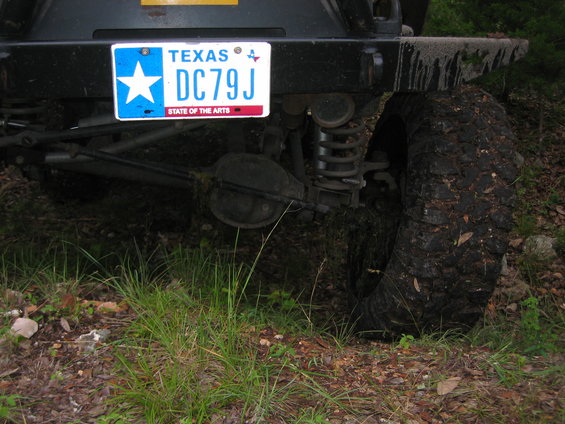 Black Jeep Wrangler rear axle and tire covered in mud, with a Texas license plate