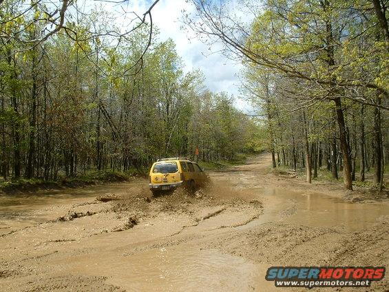 Yellow SUV driving through a muddy forest road, kicking up mud