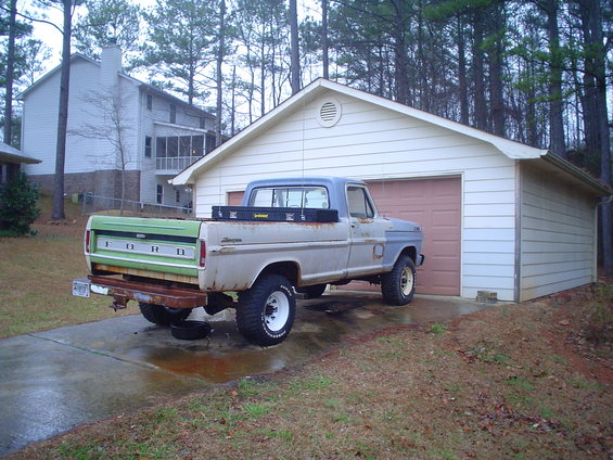 Gray and green 1967 Ford F-250 pickup truck parked in front of a garage, showing rust and wear