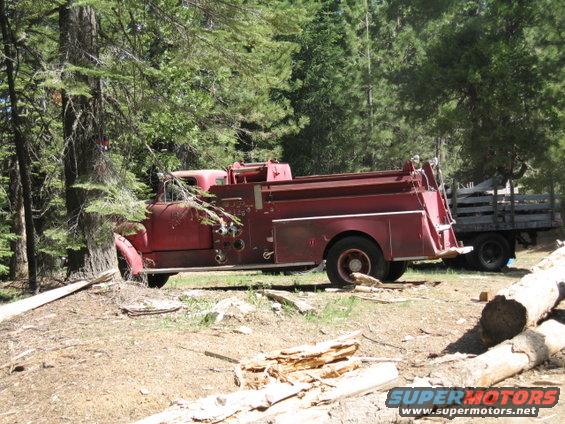 Red fire truck parked in woods, side view. Another truck with wooden bed behind it
