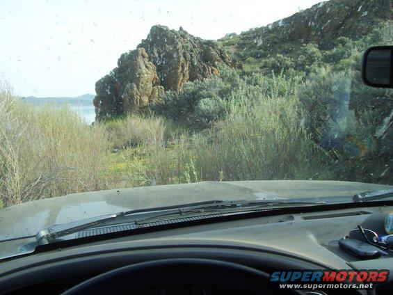 View from inside a vehicle looking out at a rocky hill and some bushes. There's a lake in the distance