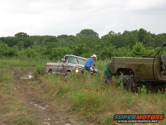 Two trucks stuck in a muddy field with people trying to free one