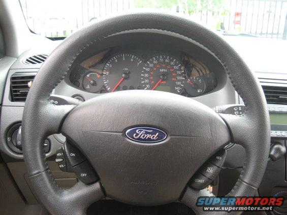 Driver's view of a Ford steering wheel and dashboard with gauges. The Ford logo is visible on the steering wheel