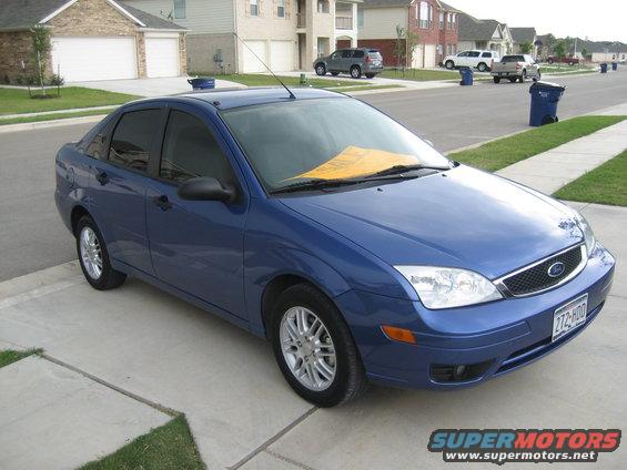 Blue sedan parked on driveway, yellow paper on windshield. Houses and other cars in background