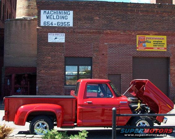 Red pickup truck with hood open, parked in front of brick building with signs