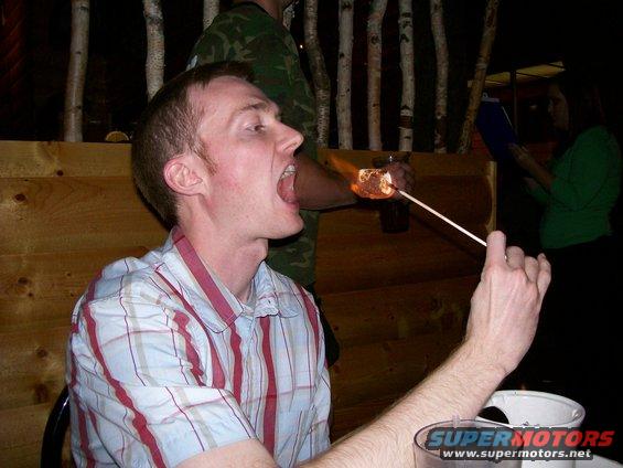Man eating a flaming marshmallow over a campfire setting