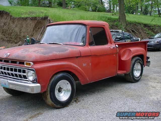 Red 1965 Ford F-100 pickup truck with chrome grille and aftermarket wheels, parked on gravel