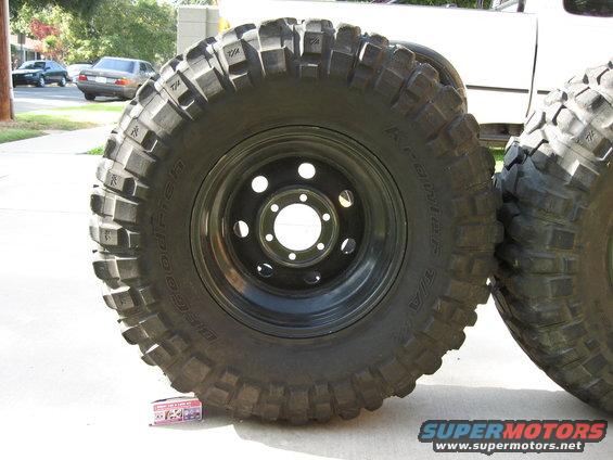 BFGoodrich off-road tires mounted on black rims, one tire is leaning on another. A white truck is partially visible in the background